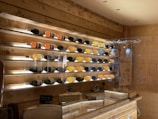 A wooden display wall featuring several rows of wine bottles, each with different colored liquid, illuminated by soft white lighting. The wall has a rustic style with wooden panels, and there are chalkboards with writing below the display. A countertop is visible in the foreground, suggesting a cozy wine bar or shop setting.