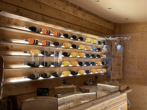 A wooden display wall featuring several rows of wine bottles, each with different colored liquid, illuminated by soft white lighting. The wall has a rustic style with wooden panels, and there are chalkboards with writing below the display. A countertop is visible in the foreground, suggesting a cozy wine bar or shop setting.