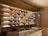 A wooden display wall featuring several rows of wine bottles, each with different colored liquid, illuminated by soft white lighting. The wall has a rustic style with wooden panels, and there are chalkboards with writing below the display. A countertop is visible in the foreground, suggesting a cozy wine bar or shop setting.