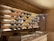 A wooden display wall featuring several rows of wine bottles, each with different colored liquid, illuminated by soft white lighting. The wall has a rustic style with wooden panels, and there are chalkboards with writing below the display. A countertop is visible in the foreground, suggesting a cozy wine bar or shop setting.