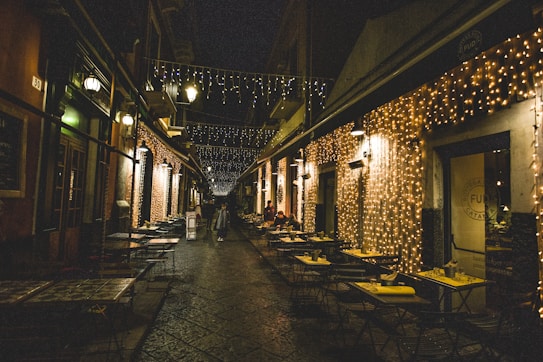 A narrow cobblestone street is lined with small caf&eacute; tables on both sides. The buildings are adorned with twinkling string lights, creating a warm and inviting atmosphere. A few people are seen walking and sitting, suggesting a relaxed evening setting.