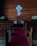 A small church interior with wooden pews, a red carpet leading to a pulpit with a cross engraved on it, and a neon blue illuminated cross on the wooden paneled wall behind the pulpit. Two sets of candles are placed on stands on either side.