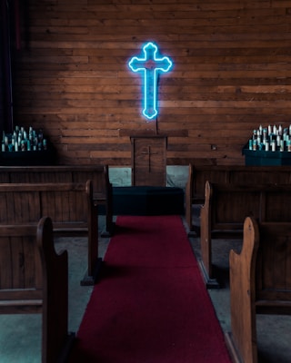 A small church interior with wooden pews, a red carpet leading to a pulpit with a cross engraved on it, and a neon blue illuminated cross on the wooden paneled wall behind the pulpit. Two sets of candles are placed on stands on either side.