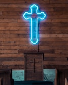 A neon blue cross is mounted on a wooden wall, above a wooden pulpit, which also has a carved cross on its front. The setting suggests an indoor religious space with wooden pews visible.