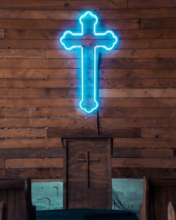 A neon blue cross is mounted on a wooden wall, above a wooden pulpit, which also has a carved cross on its front. The setting suggests an indoor religious space with wooden pews visible.