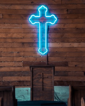 A neon blue cross is mounted on a wooden wall, above a wooden pulpit, which also has a carved cross on its front. The setting suggests an indoor religious space with wooden pews visible.
