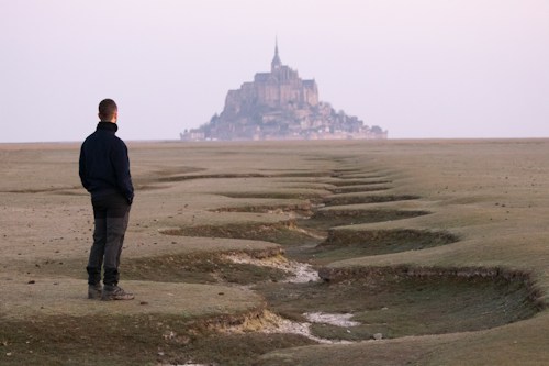A person stands on a vast, barren landscape with a winding path leading to a distant castle-like structure on a hill. The scene is serene, with muted colors and an expansive view.