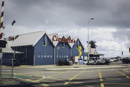 A coastal building with a blue facade and a large red-lettered sign that reads 'Cicerello's'. Multiple flags are mounted on poles atop the building, and there is a distinctive yellow sculpture near the entrance. Several cars are parked nearby, and there is a cloudy sky overhead.
