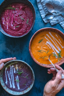 Three bowls of creamy soups, each with a different vibrant color, are arranged artfully on a dark blue surface. The top left bowl contains red soup garnished with rosemary and a drizzle of cream, the top right bowl holds orange soup with similar garnishes, while the bottom left bowl features purple soup. A folded blue napkin with two metal spoons rests beside the bowls, and human hands are seen holding one of the bowls and a spoon poised to scoop up the soup.
