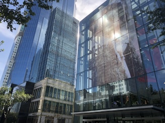 High-rise modern glass buildings reflect the sunlight, with one displaying a large advertisement on its facade. The surrounding area includes some green trees, adding contrast to the gray and blue structures.