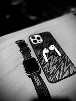 A close-up view of a black smartwatch with a UAG strap, a smartphone with a patterned case, and a pair of wireless earbuds placed on a fabric surface. The image is in black and white, emphasizing the textures and shapes of the objects.