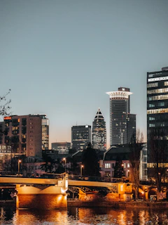 A modern city skyline at dusk with illuminated buildings reflecting on a calm river.