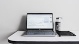 A minimalist desk setup featuring an open laptop displaying a line graph, accompanied by a water bottle with the text 'MY BOTTLE' and a closed black notebook. The background is a plain white wall, and the desk surface is glossy and white.
