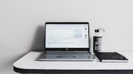 A minimalist desk setup featuring an open laptop displaying a line graph, accompanied by a water bottle with the text 'MY BOTTLE' and a closed black notebook. The background is a plain white wall, and the desk surface is glossy and white.