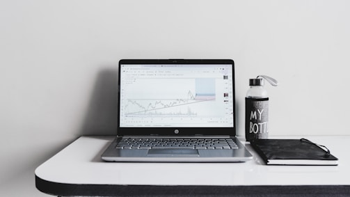 A minimalist desk setup featuring an open laptop displaying a line graph, accompanied by a water bottle with the text 'MY BOTTLE' and a closed black notebook. The background is a plain white wall, and the desk surface is glossy and white.