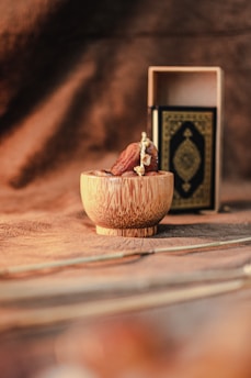 A small wooden bowl containing dates is placed on a textured brown surface. Behind it, a decorative book with a gold ornamental cover is partly visible inside an open wooden box.