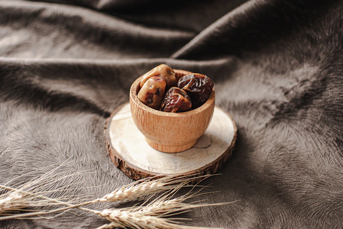 A small wooden bowl of dates set on a rustic wood slice with dried wheat sprigs
