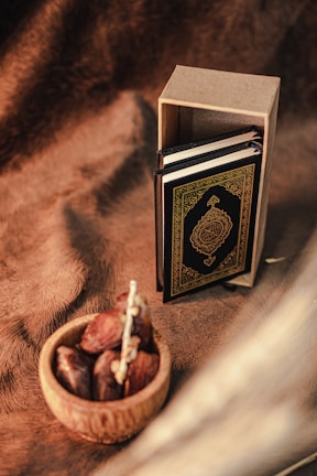 A small ornate book, possibly a Quran, is standing upright next to its cardboard box on a textured brown fabric background. In front, a wooden bowl holds several dates.
