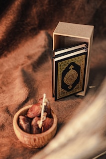 A small ornate book, possibly a Quran, is standing upright next to its cardboard box on a textured brown fabric background. In front, a wooden bowl holds several dates.