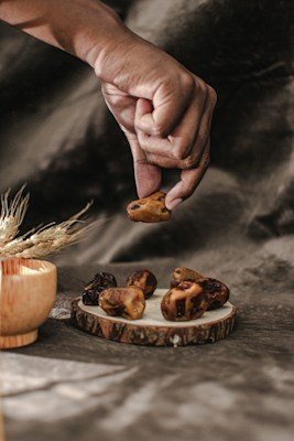 A hand is gently picking up a dried date from a rustic wooden plate with several other dates on it. The background is a soft, muted gray-brown fabric, creating a warm and cozy atmosphere. On the left, there is a wooden bowl and some decorative dried wheat stalks.