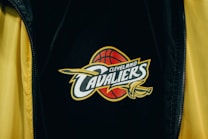A close-up view of a black garment with the Cleveland Cavaliers logo prominently displayed. The logo features a red basketball and a sword with the word 'Cavaliers' across it, emphasizing bold typography and dynamic design elements. The jacket's outer area is lined with a contrasting bright yellow fabric.