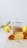 A glass teapot filled with a herbal infusion sits on a wooden surface next to a vibrant orange chrysanthemum flower. In the background, there are yellow quince fruits, partially blurred to emphasize the teapot. The image has a minimalistic and bright aesthetic.