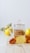 A glass teapot filled with a herbal infusion sits on a wooden surface next to a vibrant orange chrysanthemum flower. In the background, there are yellow quince fruits, partially blurred to emphasize the teapot. The image has a minimalistic and bright aesthetic.