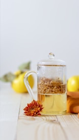 A glass teapot filled with a herbal infusion sits on a wooden surface next to a vibrant orange chrysanthemum flower. In the background, there are yellow quince fruits, partially blurred to emphasize the teapot. The image has a minimalistic and bright aesthetic.