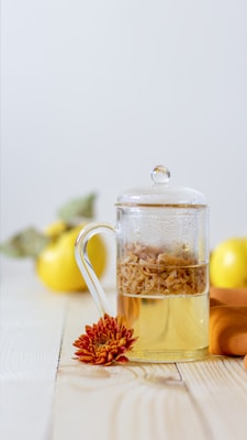 A glass teapot filled with a herbal infusion sits on a wooden surface next to a vibrant orange chrysanthemum flower. In the background, there are yellow quince fruits, partially blurred to emphasize the teapot. The image has a minimalistic and bright aesthetic.