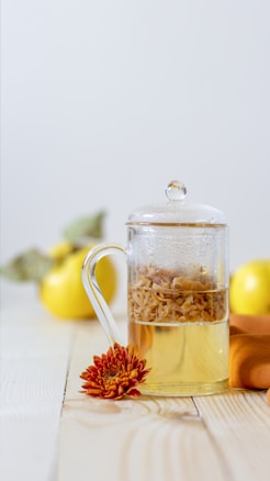A glass teapot filled with a herbal infusion sits on a wooden surface next to a vibrant orange chrysanthemum flower. In the background, there are yellow quince fruits, partially blurred to emphasize the teapot. The image has a minimalistic and bright aesthetic.