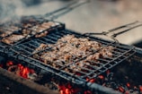 Juicy pieces of meat are sizzling on a grill, positioned over glowing charcoal embers. The scene captures the essence of a barbecue with a smoky and rustic appeal, enhanced by the visible grill marks on the meat.