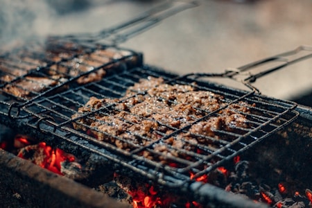 Juicy pieces of meat are sizzling on a grill, positioned over glowing charcoal embers. The scene captures the essence of a barbecue with a smoky and rustic appeal, enhanced by the visible grill marks on the meat.