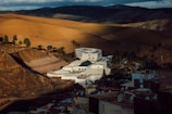 A white modern building complex with a circular section is situated amidst a mountainous landscape with vast, sunlit brown and green fields. Dark clouds loom over the hills in the background, casting shadows across the terrain. Satellite dishes are visible on rooftops in the foreground.