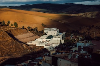 A white modern building complex with a circular section is situated amidst a mountainous landscape with vast, sunlit brown and green fields. Dark clouds loom over the hills in the background, casting shadows across the terrain. Satellite dishes are visible on rooftops in the foreground.