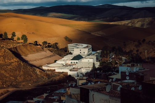 A white modern building complex with a circular section is situated amidst a mountainous landscape with vast, sunlit brown and green fields. Dark clouds loom over the hills in the background, casting shadows across the terrain. Satellite dishes are visible on rooftops in the foreground.