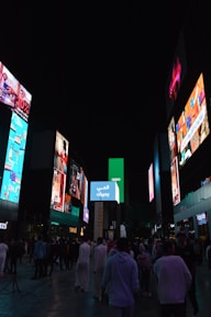 A bustling outdoor urban scene at night, featuring numerous large electronic billboards displaying advertisements. The area is crowded with people walking and socializing on a pedestrian street, and colorful screens illuminate the surroundings.