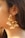 A close-up of a person's ear adorned with large, ornate gold earrings embellished with small white pearls. The earrings have a circular shape with multiple dangling pearl clusters.
