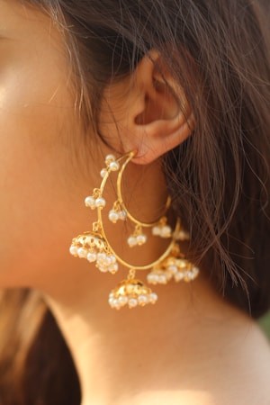 A close-up of a person's ear adorned with large, ornate gold earrings embellished with small white pearls. The earrings have a circular shape with multiple dangling pearl clusters.