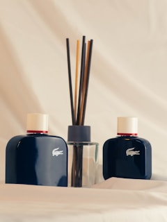 Two dark blue bottles with white caps featuring a crocodile logo are positioned on a soft, light-colored fabric background. Between the bottles is a transparent glass container fitted with several dark reed diffuser sticks.