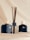 Two dark blue bottles with white caps featuring a crocodile logo are positioned on a soft, light-colored fabric background. Between the bottles is a transparent glass container fitted with several dark reed diffuser sticks.