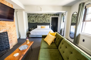 A well-lit bedroom features a large bed with white linens and a yellow decorative pillow. An olive-green sofa with patterned yellow cushions complements the interior. A wooden coffee table holds a drink with a straw and a geometric decorative piece. The room includes a brick fireplace, a flat-screen TV mounted on the wall, and a full-length mirror on the side. The wallpaper behind the bed has a leafy design, and the windows are draped with neutral curtains.