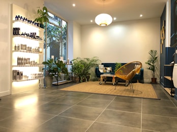 A modern, cozy living space with a minimalist aesthetic. The room features a blue velvet sofa adorned with geometric patterned cushions and a woven chair. A variety of green potted plants are placed throughout, adding a touch of nature. On the left, shelves display a range of neatly arranged bottles and products, likely skincare or haircare items. The floor is covered with large grey tiles, and a beige woven rug anchors the seating area. Warm, ambient lighting creates a comfortable atmosphere.