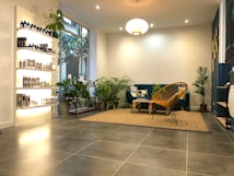 A modern, cozy living space with a minimalist aesthetic. The room features a blue velvet sofa adorned with geometric patterned cushions and a woven chair. A variety of green potted plants are placed throughout, adding a touch of nature. On the left, shelves display a range of neatly arranged bottles and products, likely skincare or haircare items. The floor is covered with large grey tiles, and a beige woven rug anchors the seating area. Warm, ambient lighting creates a comfortable atmosphere.