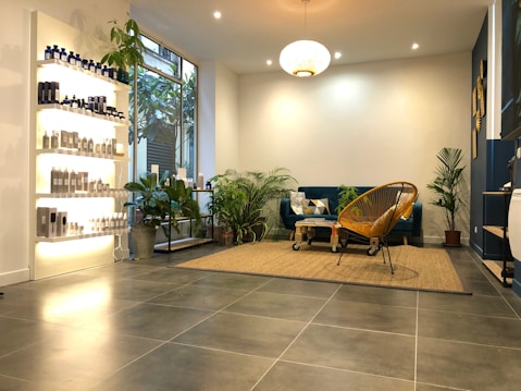 A modern, cozy living space with a minimalist aesthetic. The room features a blue velvet sofa adorned with geometric patterned cushions and a woven chair. A variety of green potted plants are placed throughout, adding a touch of nature. On the left, shelves display a range of neatly arranged bottles and products, likely skincare or haircare items. The floor is covered with large grey tiles, and a beige woven rug anchors the seating area. Warm, ambient lighting creates a comfortable atmosphere.