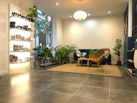 A modern, cozy living space with a minimalist aesthetic. The room features a blue velvet sofa adorned with geometric patterned cushions and a woven chair. A variety of green potted plants are placed throughout, adding a touch of nature. On the left, shelves display a range of neatly arranged bottles and products, likely skincare or haircare items. The floor is covered with large grey tiles, and a beige woven rug anchors the seating area. Warm, ambient lighting creates a comfortable atmosphere.