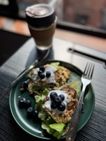 A green plate holds two pancakes topped with yogurt and fresh blueberries, placed on a bed of lettuce. A stainless steel fork and knife are positioned beside the plate. In the background, a cup with a lid appears to be filled with a beverage.