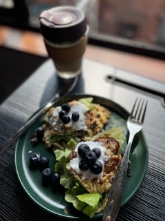 A green plate holds two pancakes topped with yogurt and fresh blueberries, placed on a bed of lettuce. A stainless steel fork and knife are positioned beside the plate. In the background, a cup with a lid appears to be filled with a beverage.