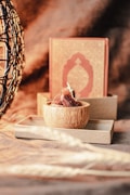 A wooden bowl filled with dates is placed on a rectangular tray, with an ornate book with an intricate design in the background. To the left, a woven structure with an abstract pattern adds texture to the scene. The lighting is warm and casts soft shadows, creating an inviting atmosphere.