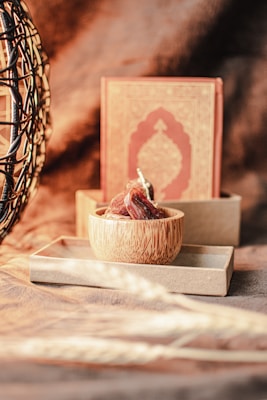 A wooden bowl filled with dates is placed on a rectangular tray, with an ornate book with an intricate design in the background. To the left, a woven structure with an abstract pattern adds texture to the scene. The lighting is warm and casts soft shadows, creating an inviting atmosphere.