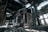 An abandoned industrial building with large, hollow structures and broken walls surrounded by rubble on the ground. Sunlight streams through the partially collapsed roof, casting dramatic shadows and highlighting the decaying architecture.
